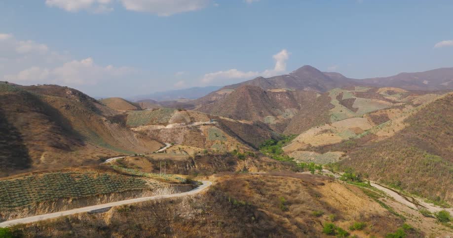 An aerial wide view of a vast dry mountain range shows a winding road and terraced hillsides planted with rows of agave plants under a blue sky with scattered white clouds
