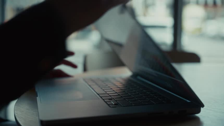 A persons hands operate a laptop computer on a table near a window before the lid is lowered and closed ending the activity