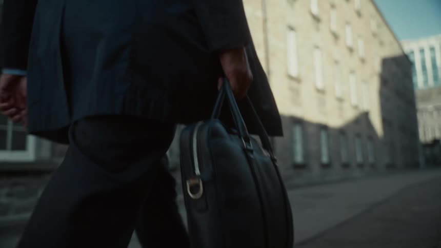 The video shows a closeup lowangle shot of a person dressed in a dark suit or professional attire walking along an urban street they are carrying a black leather briefcase with silver hardware in their right hand with a large lightcolored brick bui