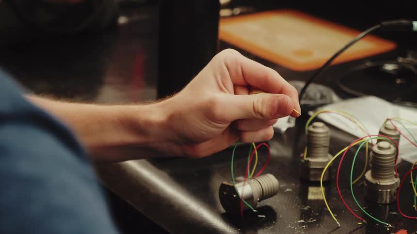 A persons hands wire a small electric motor at a cluttered workstation with red wire strippers nearby