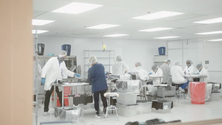 Several people in white clean room suits masks and hair nets work on products at tables in a brightly lit industrial environment