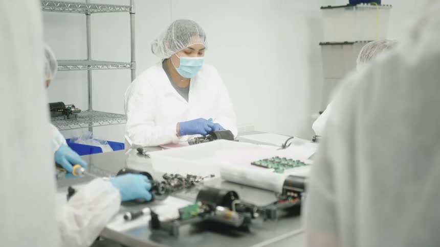 A medium shot shows three people wearing white fullbody cleanroom suits and face masks while working at a table in a sterilelooking environment they are all focused intently on small electronic or mechanical components spread across the white s