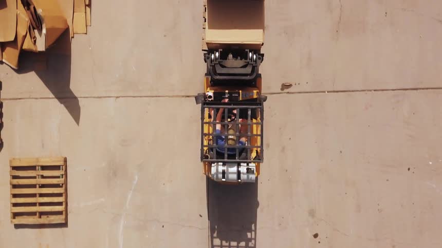 The video is a direct overhead aerial shot of a forklift maneuvering on a large flat concrete or asphalt yard casting a long shadow the bright yellow forklift is carrying a wooden pallet and passes near other pallets visible on the perimeter
