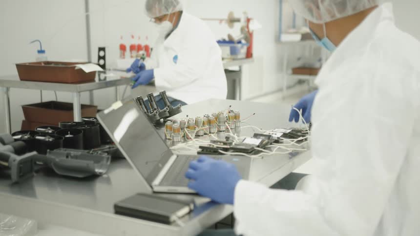 A medium view of two workers in a bright clean room or laboratory wearing white lab coats blue gloves and face masks as they assemble small electronic parts on a metal table with an open laptop in the foreground