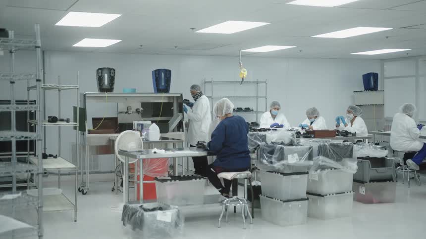 People wearing white protective suits hair nets and masks work at tables in a brightly lit clean room assembling or inspecting electronic equipment