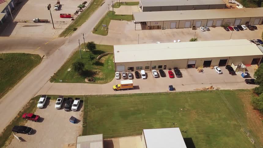 An aerial view shows a small industrial park with several beige warehouse buildings and a central parking lot filled with various cars and trucks on a sunny day