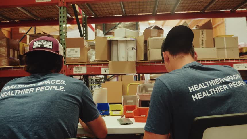 A medium rear view shows two men wearing dark blue tshirts with healthier spaces healthier people printed on the back sitting at a table working inside a warehouse lined with shelves of cardboard boxes
