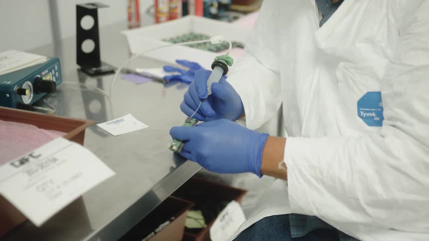 Person in white lab coat and blue gloves handling electronic component at technical workstation