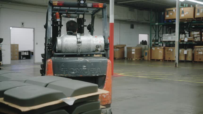 A man operates a forklft carrying a high column of round black items the vehicle moves backward away from the camera and the indoor space contains rows of stacked inventory and products on metal racks