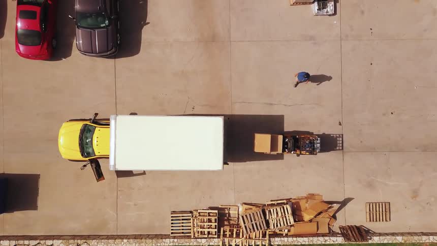 An aerial high angle shot looks directly down at a bright yellow box truck being unloaded by a worker driving a small forklift in a sunny parking lot full of parked cars and scattered wooden pallets