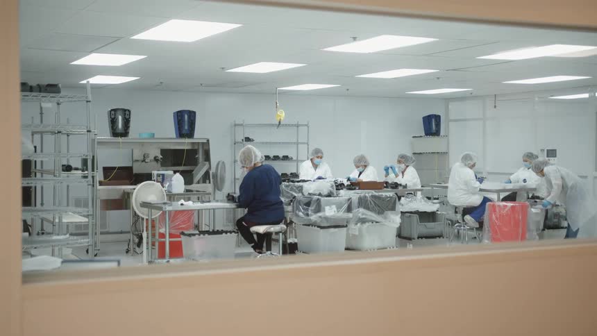 Multiple workers in protective clothing including hairnets and masks are seated at tables and engaged in precise work within a brightly lit laboratory all viewed through a window in a doorway