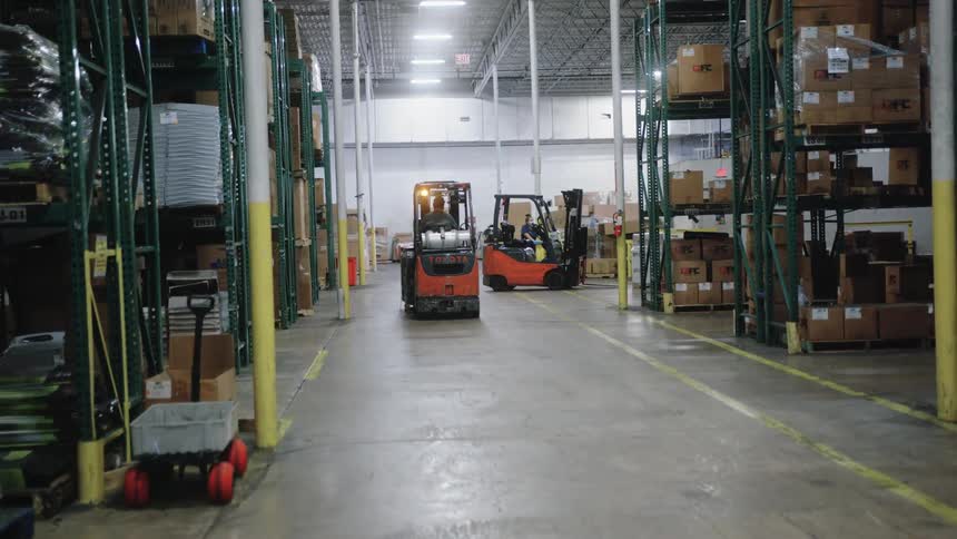 The video shows an industrial scene with a red and black toyota forklift moving through the aisles of a warehouse the area is surrounded by tall metal shelving units stacked with numerous cardboard boxes