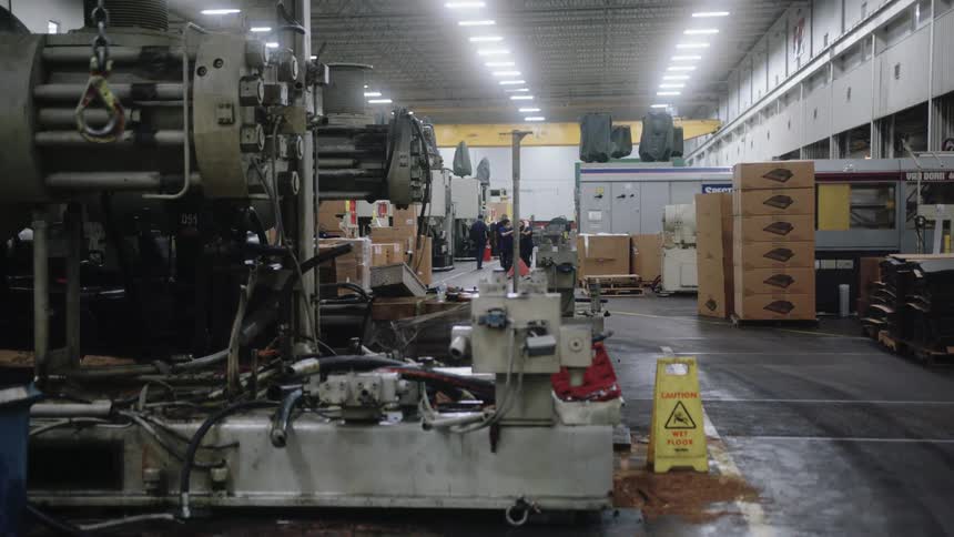A medium view inside a brightly lit factory shows a large complex piece of industrial machinery in the foreground with a group of workers gathered in the distant background near tall stacks of cardboard boxes