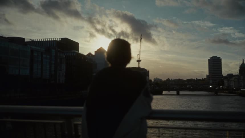 Silhouetted person stands on urban bridge with river in the background during sunset