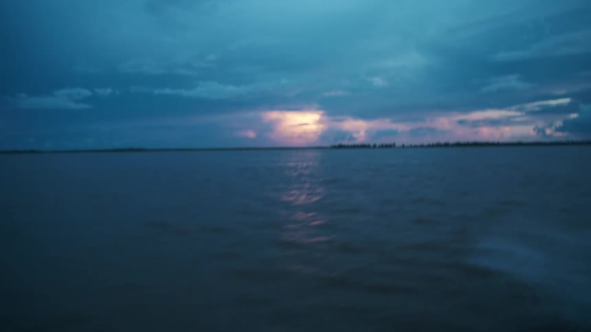 The video is an establishing shot of a wide body of water under a dark dramatic and stormy sky likely at twilight or dawn a small strip of distant land is visible on the horizon with sunlight peaking through the clouds and reflecting off