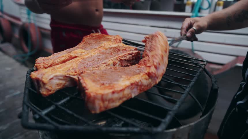 A person picking up meat with another person and placing it on a barbecue grill
