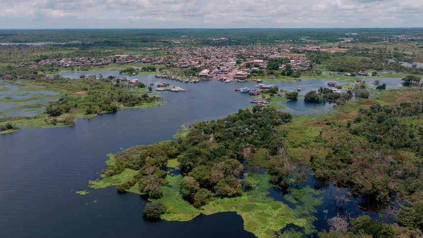 The video is a high wide aerial shot overlooking a small village surrounded by dense lush green forest and swampy dark waters suggesting a remote location like the amazon basin numerous small buildings and homes many appearing to be built on or