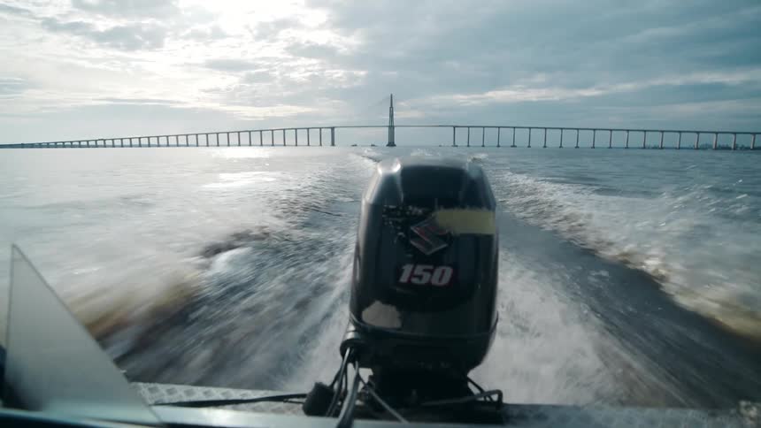 A low rear view from a fast moving boat shows a 150 horsepower outboard motor churning a white wake across a wide body of water towards a long distant bridge under an overcast sky
