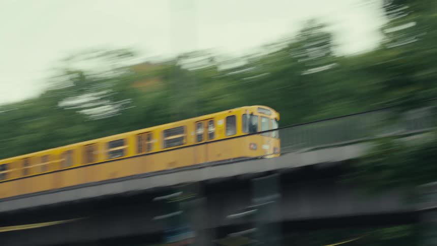 A motion blurred side medium view captures a fast moving yellow subway train traveling quickly across a gray elevated bridge with bright green trees and buildings in the background (slow shutter)
