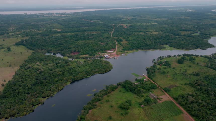 The video provides a high sweeping aerial view of a winding dark river surrounded by dense green forestin the center of the frame there is a small clearing with a few structures and a dirt road running near the riverbank connecting to the b