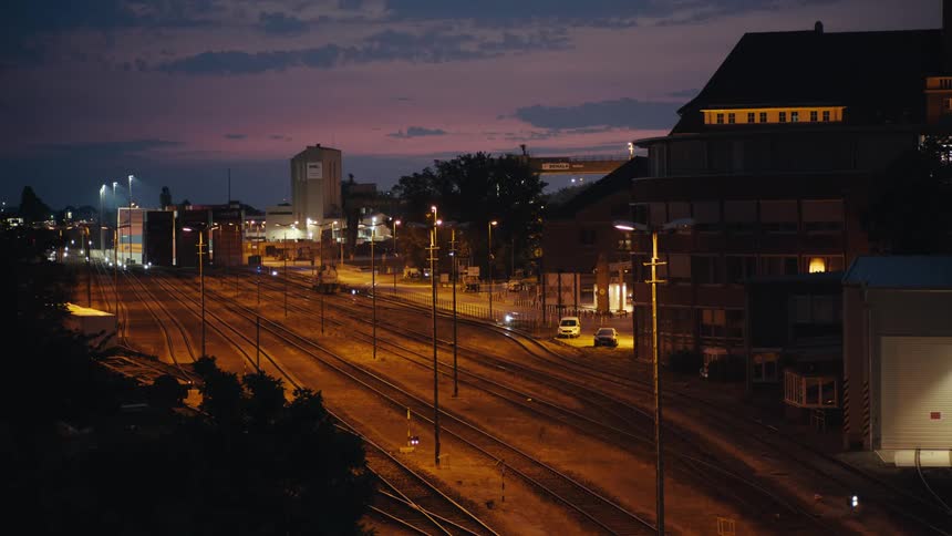 Numerous metal rails cross a level ground area a row of lit vertical poles separates the tracks dark buildings stand on the periphery and the atmosphere is illuminated by artificial light under a fading evening sky