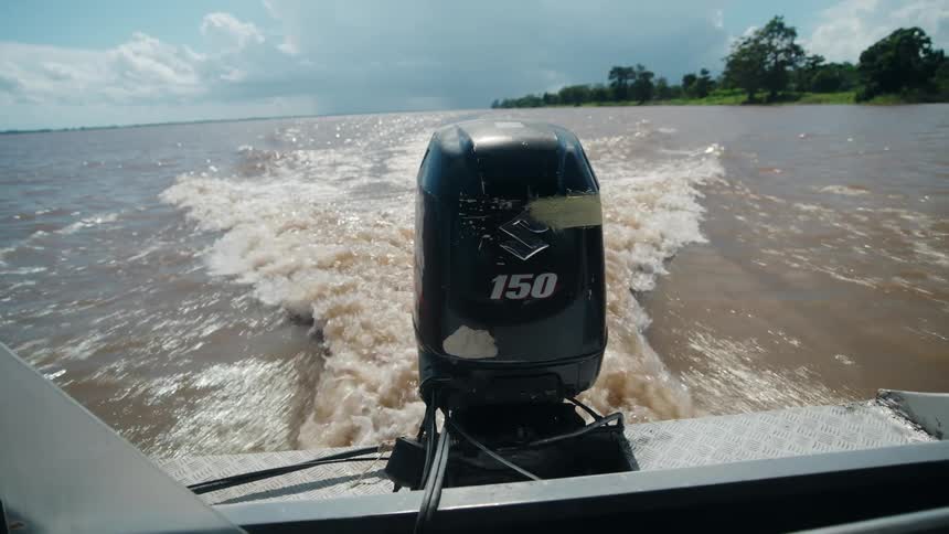 A low angle view from the rear of a moving boat shows the black 150 horsepower outboard motor churning brown river water leaving a white wake with a green shoreline in the distance under a partly cloudy sky