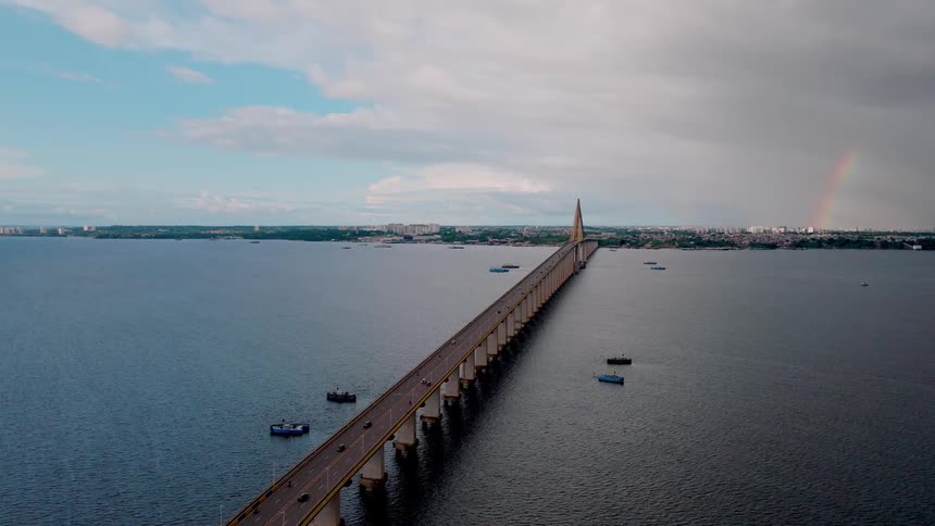 An aerial shot captures a long cablestayed highway bridge stretching far over a large body of water toward a distant city skyline under a dramatic cloudy sky a subtle rainbow arch is visible in the clouds near the city adding a striking
