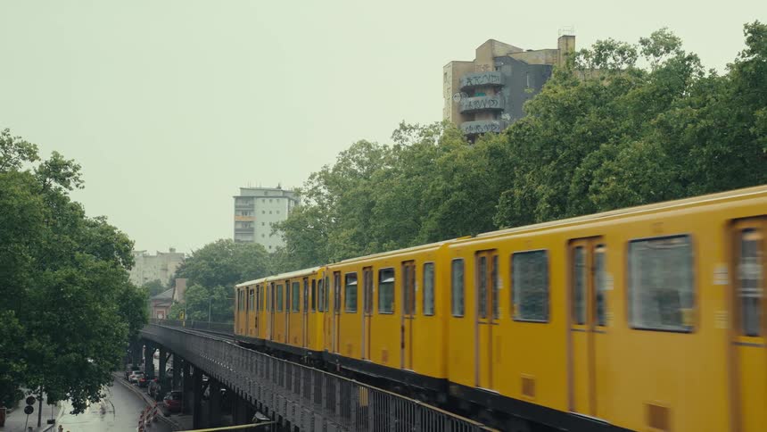 A yellow subway train moves quickly along elevated tracks passing between tall green trees apartment buildings visible in the background the sky a uniform gray