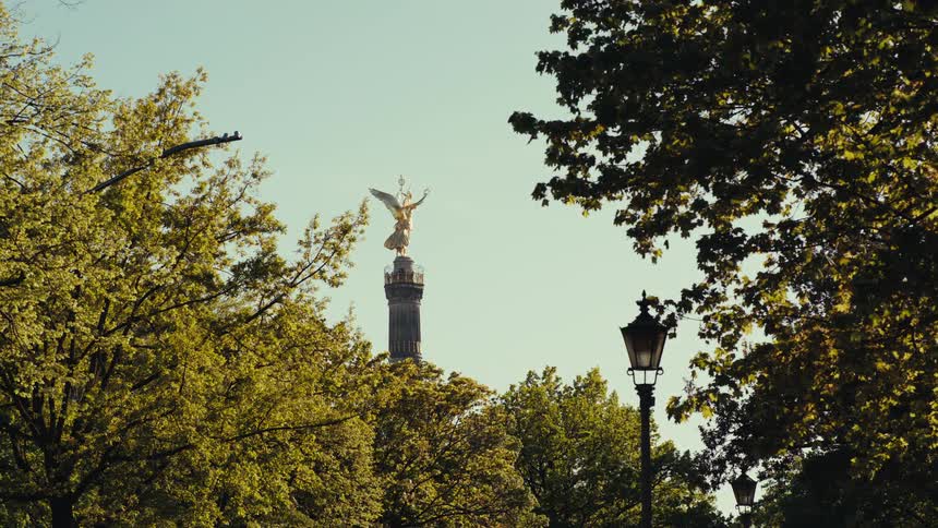 A long view shows the golden victory column statue in berlin rising above the bright green trees framed by dark foliage and an old street lamp against a clear sky