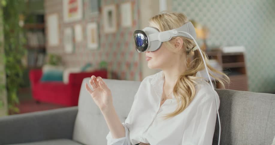 A young woman wears a vr headset device over the eyes moves a hand in the air in a gesture of control a cord extends from the back of the equipment and the background contains household items and wall decorations