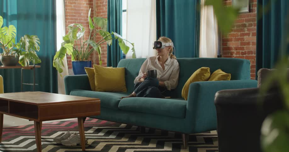 A camera shot shows a woman sitting on a teal sofa in an indoor living space with a brick wall and plants wearing a virtual reality headset and adjusting it with her hand