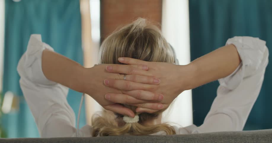A rear close view of a blonde woman wearing a grey vr goggles with cables attached as she stretches and clasps her hands behind her head over the back of a couch