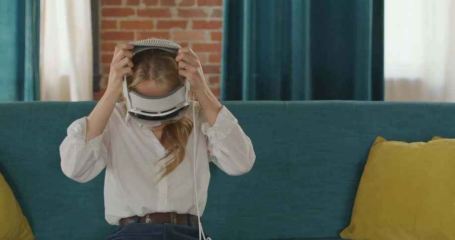 A young blond woman wearing a white shirt and jeans picks up a virtual reality headset and controller from a wood table before sitting on a teal couch and putting the headset over her eyes
