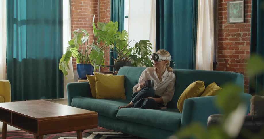 A young woman sitting on a couch in the living room wearing a virtual reality headset moving her hands and getting startled by something she saw