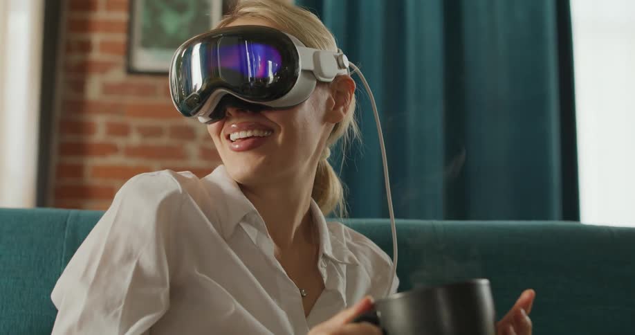 A blond woman wearing a white shirt and a sleek virtual reality headset with mirrored lenses smiles broadly while holding a black mug of hot steaming liquid on a teal sofa