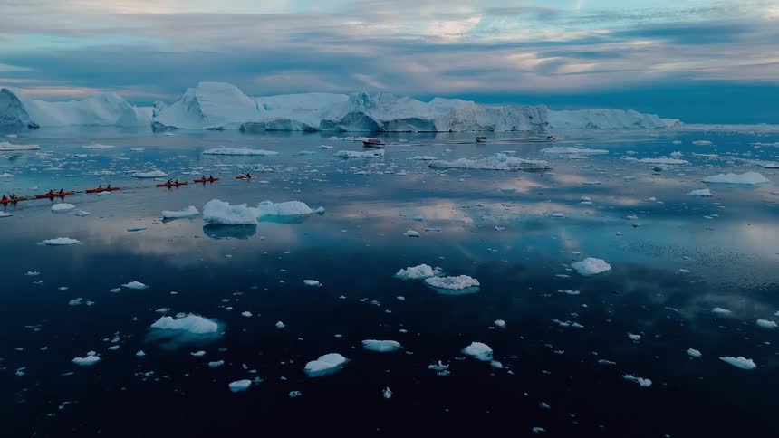 An aerial camera shot moves forward over a dark water body scattered with small ice chunks showing a line of kayaks and motorboats moving near a barrier of immense icebergs under a clouded sky