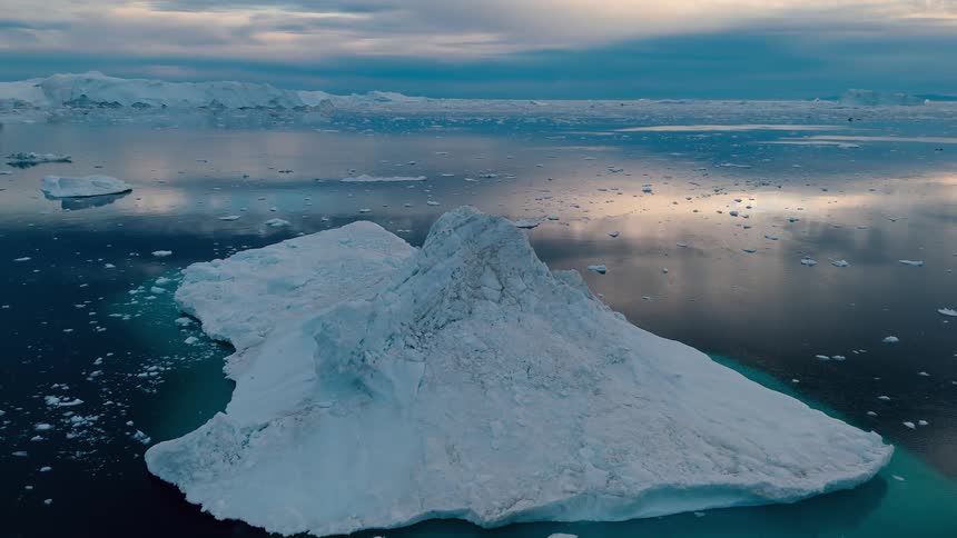 An aerial high view slowly rotates over a massive rugged white iceberg floating in dark blue water with other large ice formations visible in the distance under a dramatic cloudy sky