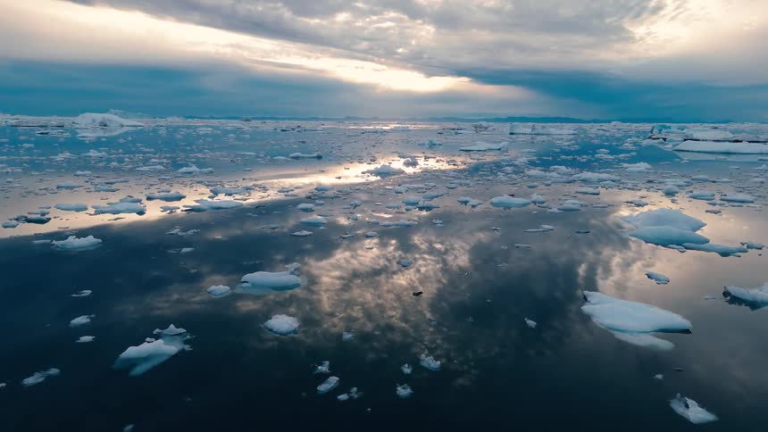 An aerial view flies smoothly over a vast calm sea filled with countless ice floes the dark water perfectly reflecting the dramatic cloudy sky