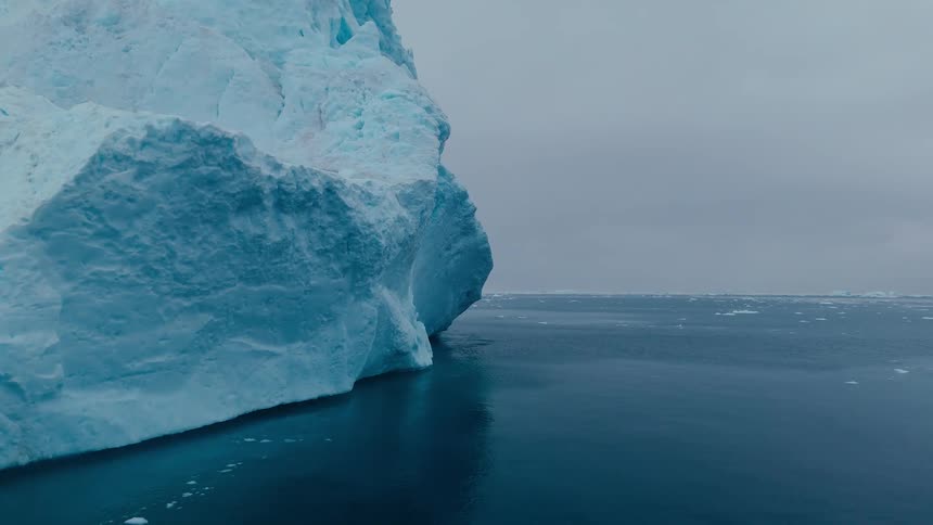 A large vertical mass of bluetinged frozen water occupies the left side the base of the structure meets the dark sea the water surface shows a deep reflection and a gray sky extends over the water and distant ice chunks