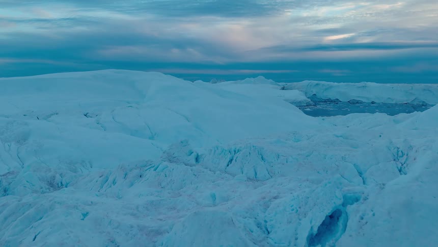 A massive textured wall of bluewhite ice rises from the dark water filling the foreground as the camera slowly rises revealing an expanse of jagged ice formations under a cloudy twilight sky