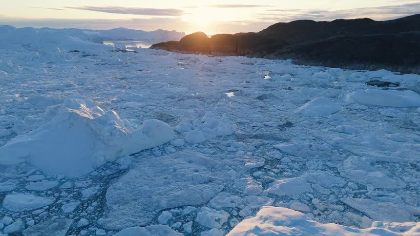 An aerial shot glides over a massive field of icebergs and broken sea ice in a polar region with dark rocky land and the setting sun on the horizon