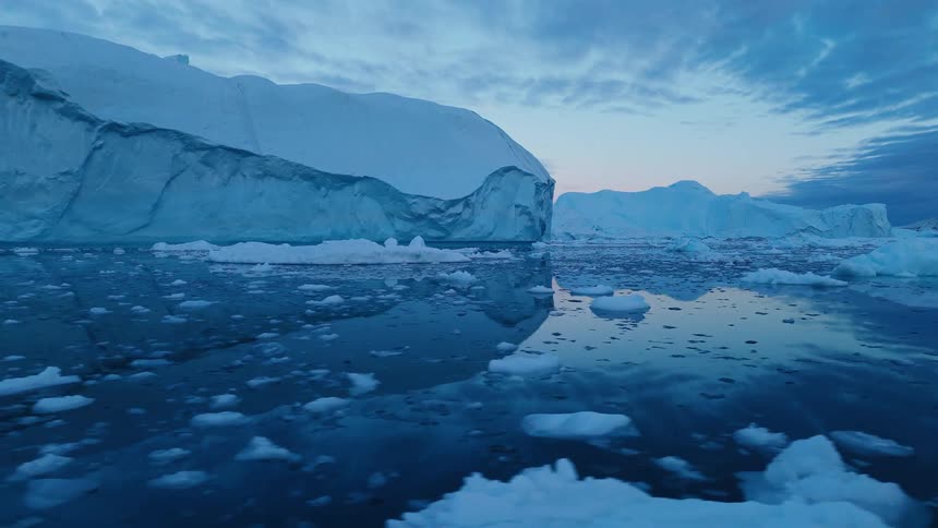 Gigantic blue and white icebergs dominate a dark cold ocean at twilight with the waters surface reflecting the dramatic cloudy blue sky