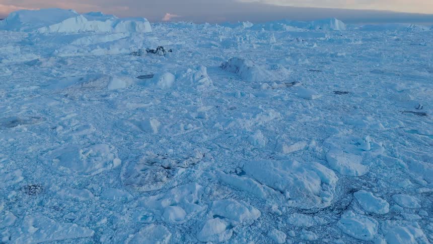 An aerial view of the arctic sea shows a vast field of broken ice floes and glacial fragments with small patches of blue water under a colorful twilight sky