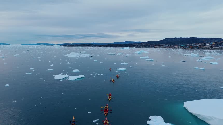 A group of kayakers paddles through a vast expanse of icy water weaving through numerous icebergs with a distant coastal town visible under a cloudy sky