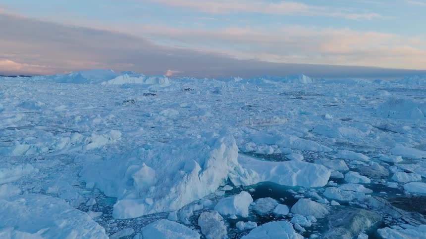 An aerial wide view shows a vast dense field of large blue and white icebergs and broken sea ice floating on the ocean under a pale colorful sky at sunset