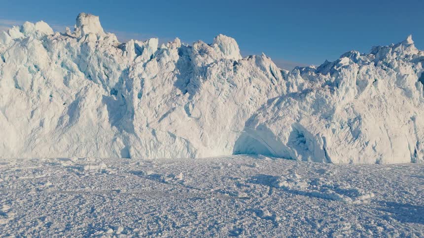 A vast towering glacier wall riddled with crevices and showing hints of blue ice rises dramatically from a field of fractured sea ice under a bright blue sky