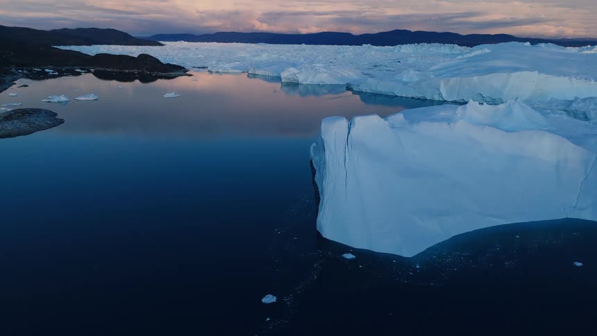 An aerial shot captures the high sheer blue and white ice cliffs of a glacier dropping sharply into a dark calm fjord the background features more floating ice and rocky snowdusted mountains under a pale evening sky creating a stark icy lan