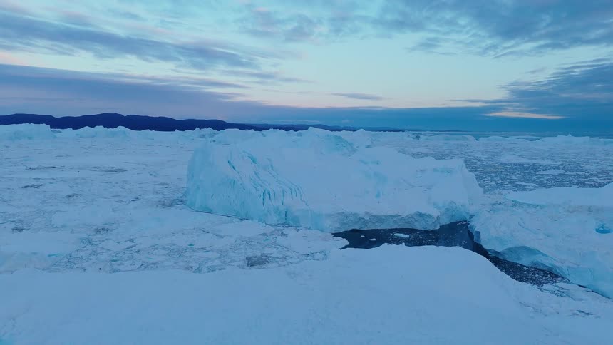 The video is an aerial shot of a vast frozen landscape covered in bright textured blue and white ice in the distance a range of dark mountains meets a pale blue and pink twilight sky contrasting with the icy foreground