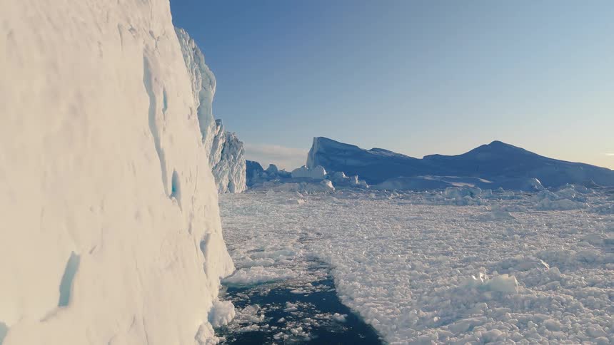 A sunlit wall of ice fills the left foreground while a field of broken sea ice leads toward distant blue bergs under a clear sky