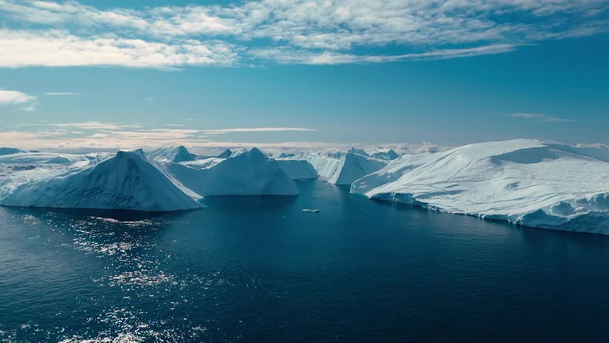 The video is a sweeping aerial shot of a large glacial fjord under a bright blue and white sky massive bright white and blue icebergs float in the deep blue water surrounded by towering snowcovered landmasses